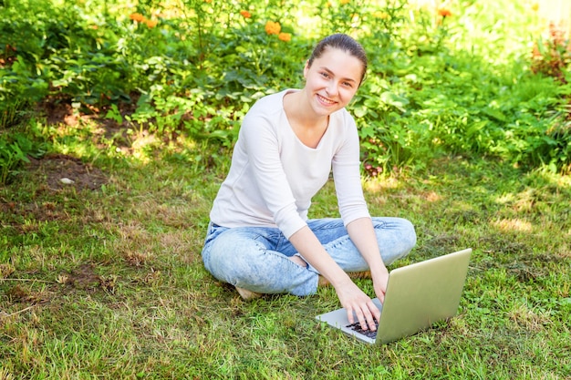 Concetto di business freelance. Giovane donna seduta sul prato di erba verde nel parco cittadino che lavora su un computer portatile. Stile di vita autentica studentessa candida che studia all'aperto. Ufficio Mobile