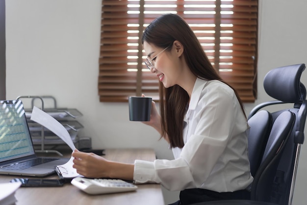 Concetto di business La donna d'affari sta leggendo un documento finanziario e sta bevendo caffè in un ufficio moderno