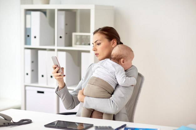 concetto di business, maternità, multi-tasking, famiglia e persone - donna d'affari sorridente con bambino e smartphone che lavora in ufficio