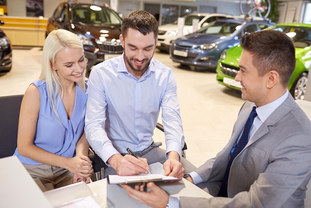 concetto di business, vendita e persone di auto - coppia felice con concessionario che acquista auto e firma documenti in auto show o salone