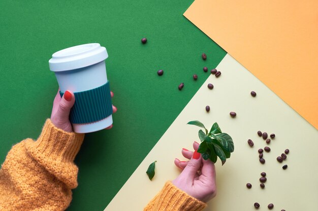 Concetto di caffè a spreco zero. Tazze di caffè riutilizzabili ecologiche nelle mani. Geometrico disteso su carta a tono diviso. Parete creativa nei colori verde, arancione e giallo.