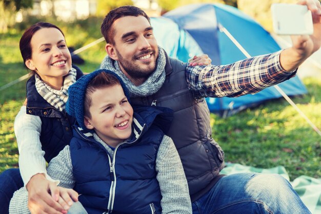 concetto di campeggio, escursione, tecnologia e persone - famiglia felice con smartphone che si fa selfie in campeggio