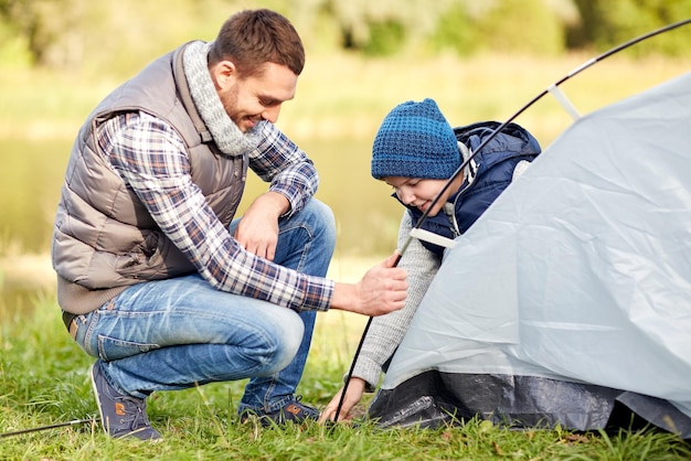 concetto di campeggio, turismo, escursione, famiglia e persone - padre e figlio felici che installano tenda all'aperto