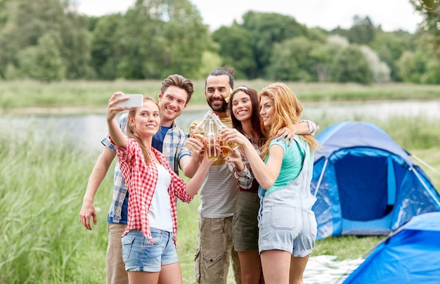 concetto di campeggio, viaggi, turismo, escursione e persone - amici felici con bottiglie di vetro che bevono sidro o birra e si fanno selfie con lo smartphone in campeggio