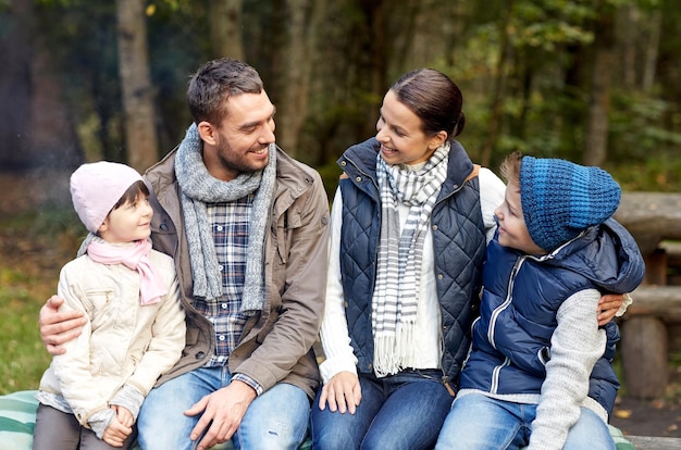 concetto di campeggio, viaggi, turismo, escursione e persone - famiglia felice seduta su una panchina e parlando al campo nei boschi