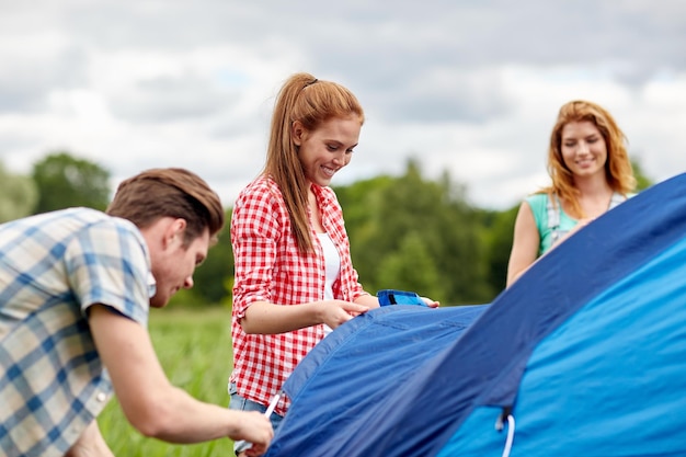 concetto di campeggio, viaggi, turismo, escursione e persone - gruppo di amici sorridenti che installano tenda all'aperto