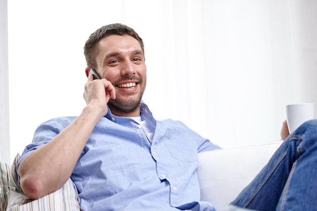 concetto di casa, tecnologia e internet - uomo sorridente con smartphone seduto sul divano di casa