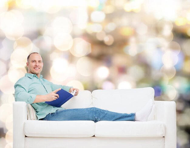 concetto di casa, tempo libero e felicità - uomo sorridente sdraiato sul divano con il libro