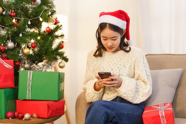 Concetto di celebrazione del Natale Giovane donna asiatica seduta in soggiorno e che chiacchiera sullo smartphone