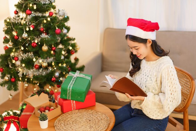 Concetto di celebrazione del Natale La giovane donna asiatica si siede su una sedia vicino all'albero di natale e legge un libro