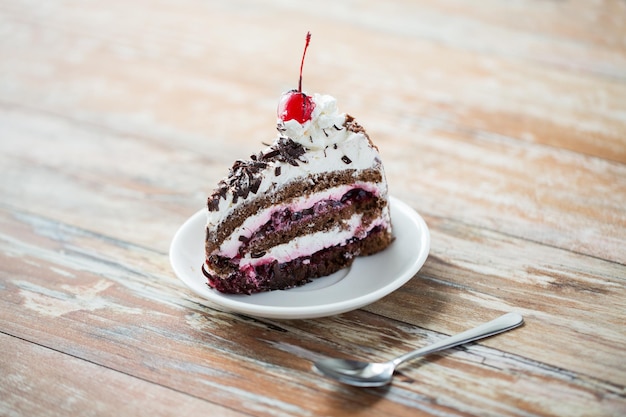 concetto di cibo, cibo spazzatura, cucina, cottura e vacanze - pezzo di deliziosa torta a strati di cioccolato ciliegia sul piattino con cucchiaio sul tavolo di legno