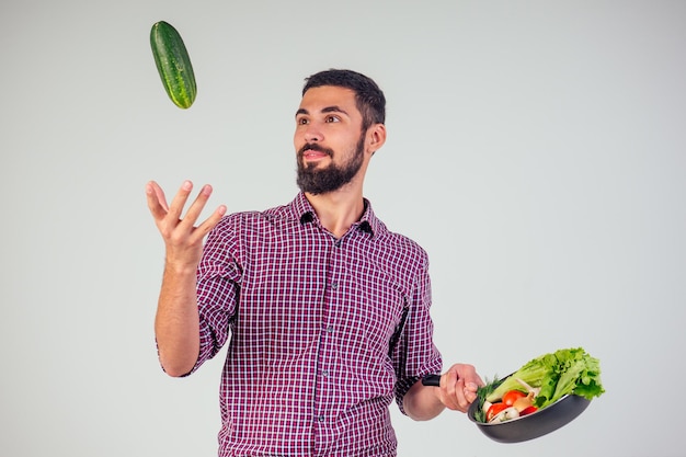 Concetto di cibo sano e vegano. Bell'uomo con la barba nera in camicia rossa a scacchi che cucina insalata con pomodori, cetriolo, funghi, cipolla e aglio sullo sfondo bianco dello studio.