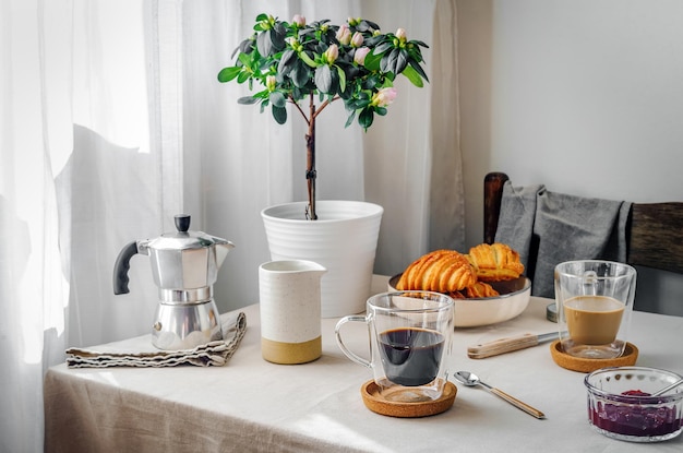 Concetto di colazione accogliente primaverile Due tazze con caffè e croissant al latte pianta con fiori crema caffettiera marmellata Luce mattutina attraverso tende tovaglia di lino