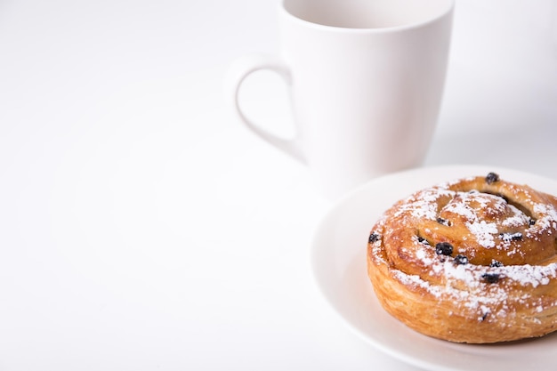 Concetto di colazione - panino dolce con uvetta e tazza di caffè o tè su sfondo bianco