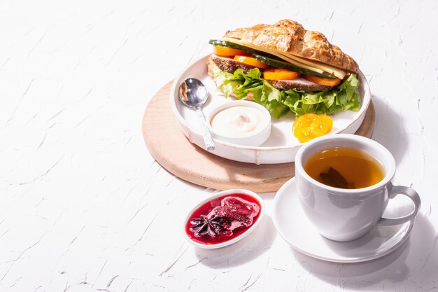 Concetto di colazione sana. Buon umore mattutino con tè, croissant e marmellata di fragole. Una luce dura alla moda, un'ombra scura, uno sfondo di stucco bianco, spazio di copia