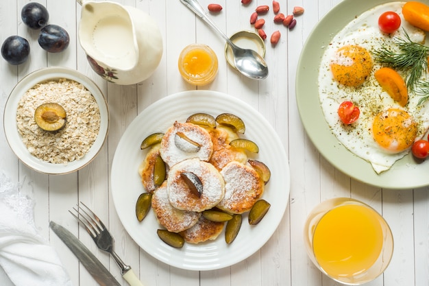 Concetto di colazione. Uova fritte, frittelle di ricotta, prugne e farina d'avena con latte, succo d'arancia sul tavolo