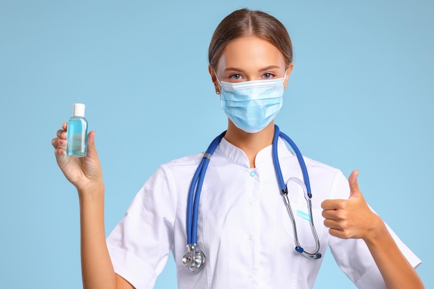 Concetto di combattere l'infezione da coronavirus un medico con una maschera medica protettiva mostra la mano disinfettante e gesticola il pollice su sfondo blu