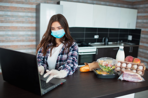 Concetto di consegna del cibo. Una giovane donna in una maschera medica e guanti di gomma usa e getta maschera ordina cibo utilizzando un computer portatile a casa. Sul tavolo ci sono latte, insalate in scatola, carne, cibo, frutta, uova, pane