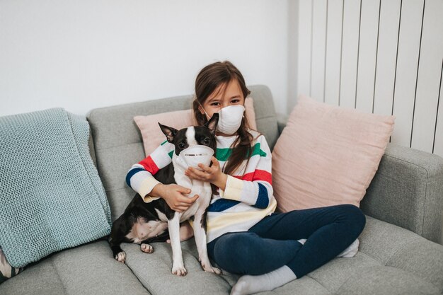 Concetto di coronavirus COVID-19. La ragazza e il suo cane con maschere protettive sul viso stanno giocando a vivere mentre sono in quarantena.