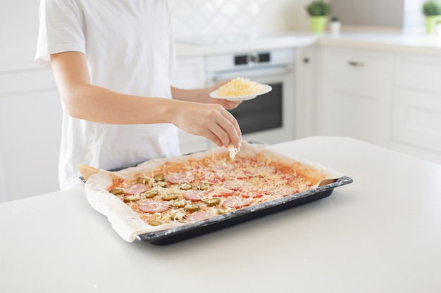 Concetto di cottura - cuocere manualmente aggiungendo formaggio grattugiato alla pizza nella cucina di casa. Avvicinamento