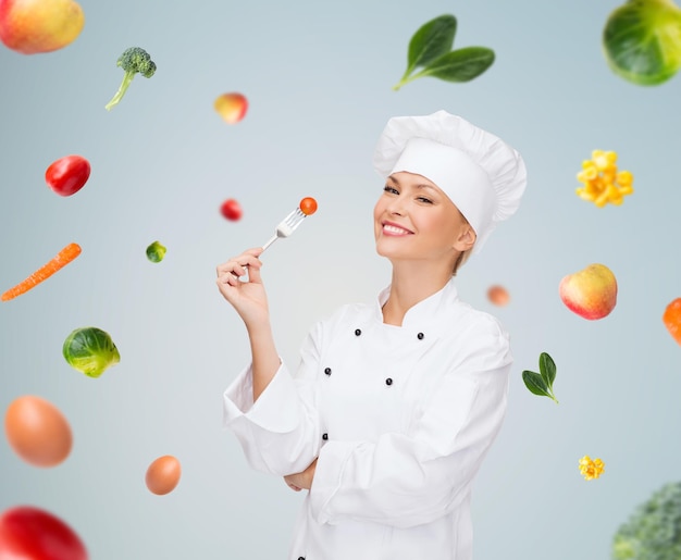 concetto di cucina e cibo - chef femminile sorridente, cuoco o fornaio con forchetta e pomodoro su verdure che cadono su sfondo grigio