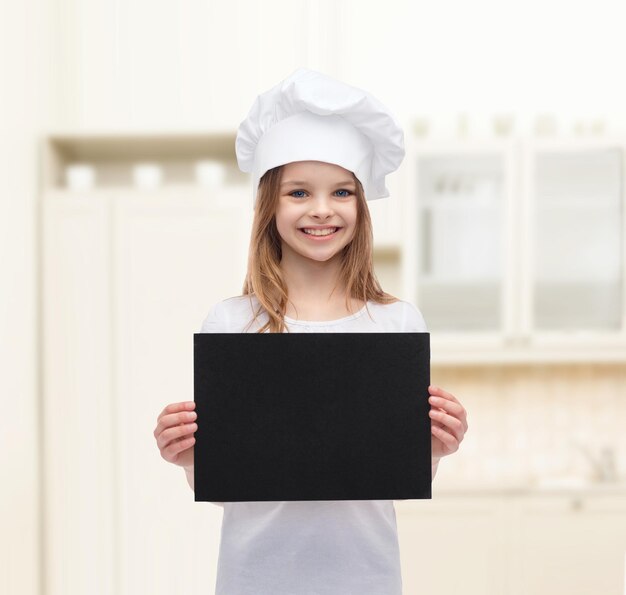 concetto di cucina, infanzia, pubblicità e persone - sorridente piccola ragazza chef, cuoco o fornaio con carta nera bianca su sfondo cucina