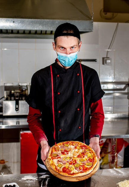 Concetto di cucina pizza, chef in maschera medica mette gli ingredienti sulla pizza