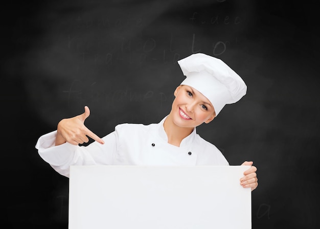 concetto di cucina, pubblicità e persone - chef femminile sorridente, cuoco o fornaio che punta il dito a bordo bianco bianco su sfondo lavagna