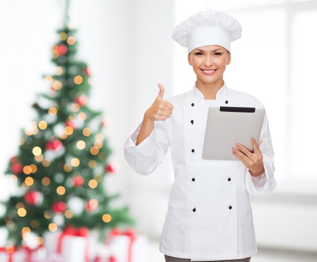 concetto di cucina, vacanze, tecnologia e persone - chef, cuoca o fornaio sorridente con computer tablet pc che mostra i pollici in su sul soggiorno con sfondo albero di natale