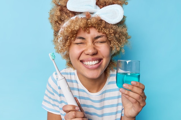 Concetto di cura orale La giovane donna riccia positiva sorride a denti stretti tiene gli occhi chiusi tiene la spazzola elettrica e il collutorio vestito casualmente si sottopone alle routine di igiene quotidiana pone su sfondo blu