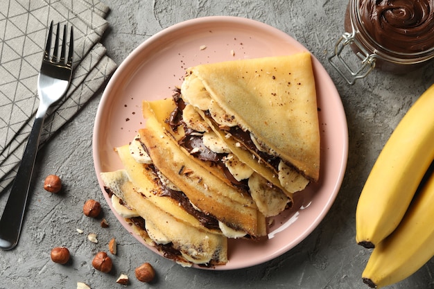 Concetto di deliziosa colazione con crepes con pasta di cioccolato, banana e noci su sfondo grigio