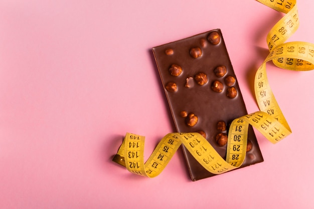 Concetto di dieta con barretta di cioccolato e nastro di misurazione per perdita di peso su sfondo rosa.