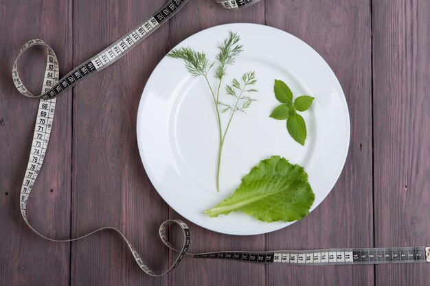 Concetto di dieta - piatto bianco con aneto, insalata di lattuga, basilico e nastro di centimetro sul tavolo di legno