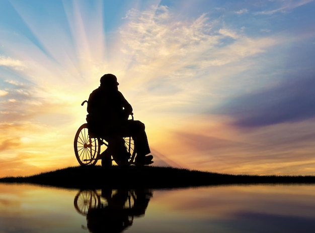 Concetto di disabilità e vecchiaia. Silhouette di persona disabile in sedia a rotelle al tramonto e riflesso nell'acqua