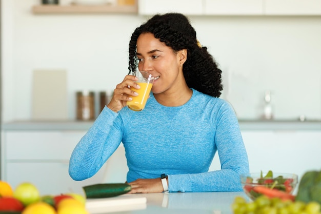 Concetto di disintossicazione Eccitata signora nera che beve succo fatto in casa seduto in cucina a preparare il pranzo dopo l'allenamento domestico