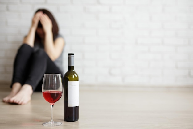 Concetto di divorzio e depressione della relazione di alcolismo femminile primo piano di vetro e bottiglia di vino e donna stressata che piange e si siede sul pavimento copia spazio sopra il muro bianco