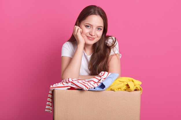 Concetto di donazione. La tenuta della donna dona la scatola con il pieno di vestiti, la femmina sorridente adorabile tiene la mano sotto il mento e i vestiti donano la scatola, signora che porta la maglietta bianca casuale, posare isolata sul rosa.