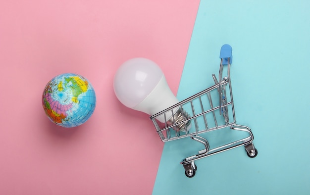 Concetto di eco. Carrello della spesa con lampadina a led e globo su fondo pastello blu-rosa. Vista dall'alto.