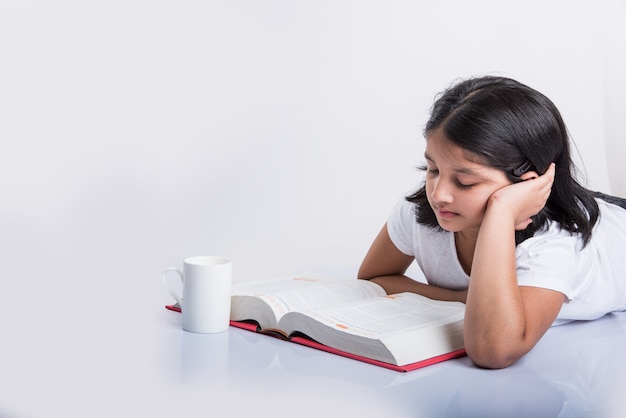 Concetto di educazione a casa - Piccola ragazza indiana o asiatica carina che studia con una pila di libri e una tazza da caffè mentre è seduta sul pavimento a casa. Isolato su sfondo bianco