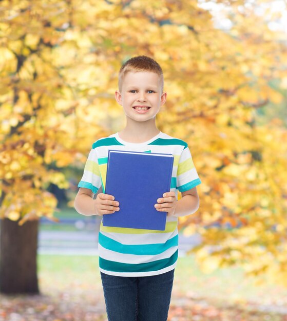 concetto di educazione, infanzia e scuola - sorridente piccolo studente ragazzo con libro blu su sfondo parco autunnale