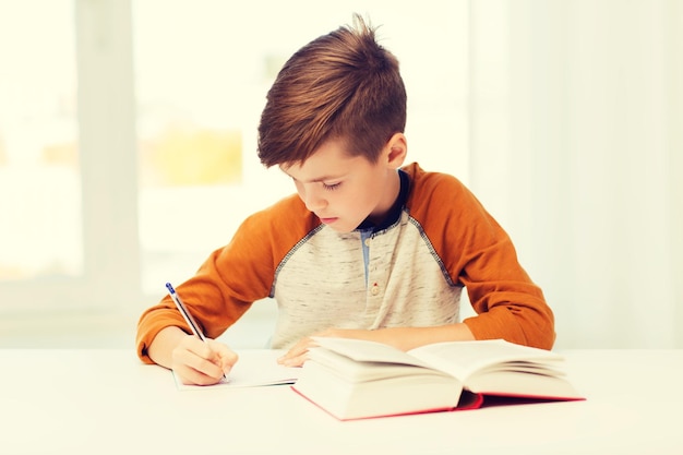 concetto di educazione, infanzia, persone, compiti a casa e scuola - ragazzo studente con libro che scrive al taccuino a casa