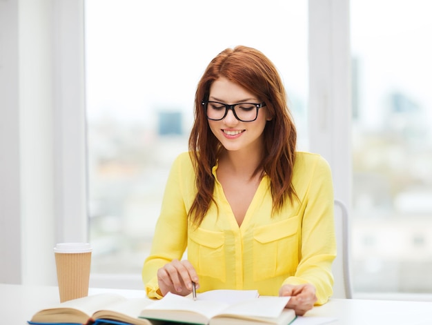 concetto di educazione - studentessa sorridente in occhiali che legge libri ana prendendo appunti in biblioteca