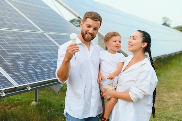 Concetto di energia solare Una giovane famiglia felice è in piedi vicino ai pannelli solari e tiene in mano una lampadina elettrica