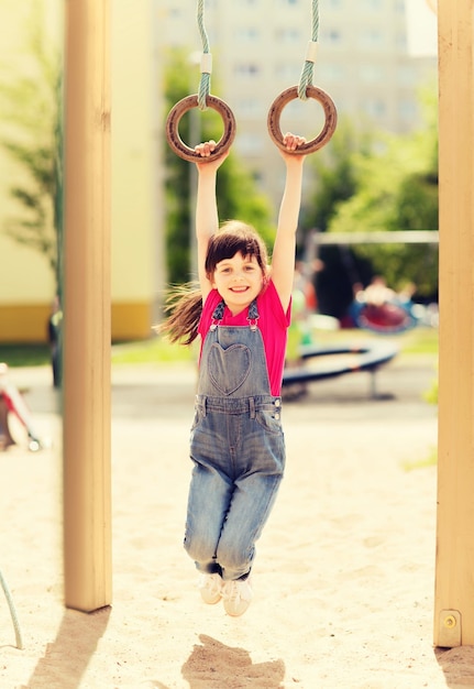 concetto di estate, infanzia, tempo libero e persone - bambina felice appesa su anelli ginnici al parco giochi per bambini