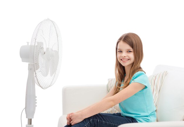 concetto di estate, tempo e attrezzatura - bambina sorridente seduta sul divano con un grande ventilatore a casa