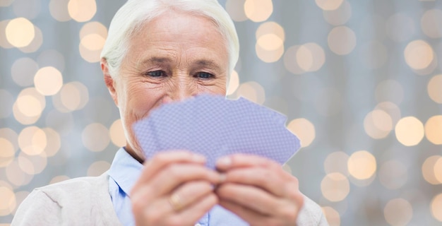 concetto di età, gioco, gioco d'azzardo, poker e persone - primo piano di una donna anziana sorridente felice che gioca a carte su sfondo di luci natalizie