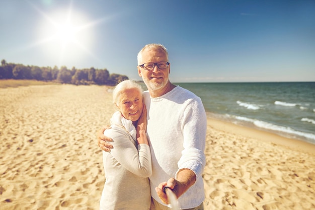 concetto di età, viaggi, turismo, tecnologia e persone - coppia senior felice che scatta foto con il selfie stick dello smartphone sulla spiaggia estiva