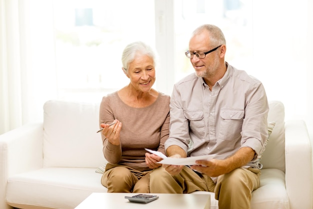 concetto di famiglia, affari, risparmio, età e persone - coppia senior sorridente con documenti e calcolatrice a casa