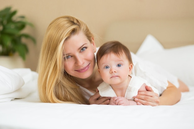 Concetto di famiglia amorevole felice. Bella madre che gioca con la sua bambina in camera da letto. Sorridono e si abbracciano insieme su lenzuola bianche