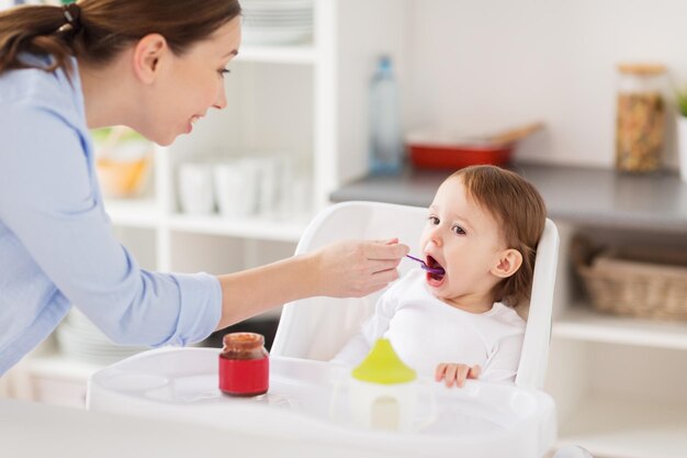 concetto di famiglia, cibo, alimentazione e persone - madre felice con purea e cucchiaio che alimenta il piccolo bambino seduto nel seggiolone a casa